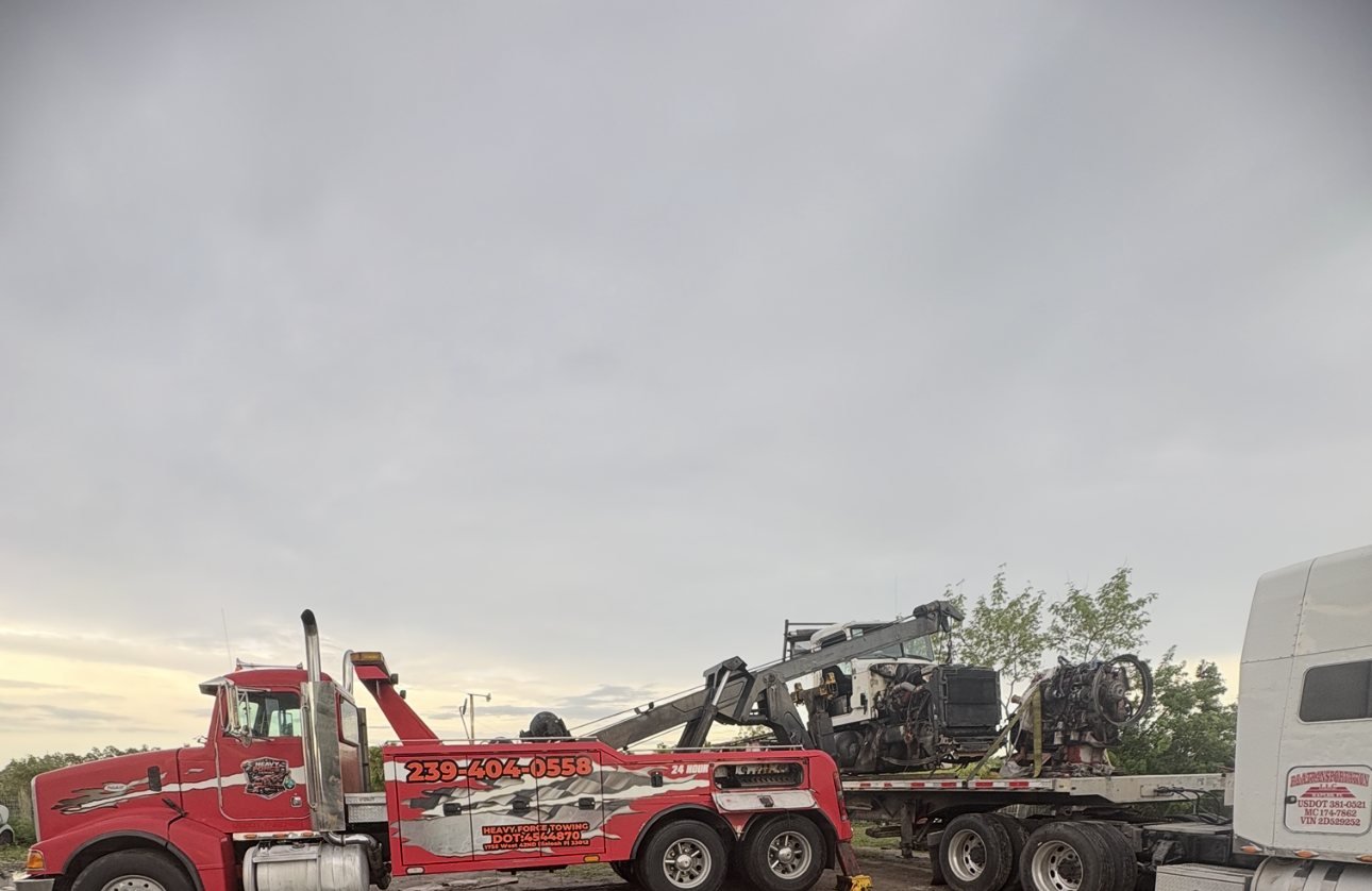 The Heavy Force wrecker at dusk beside a flatbed trailer carrying a stripped truck chassis